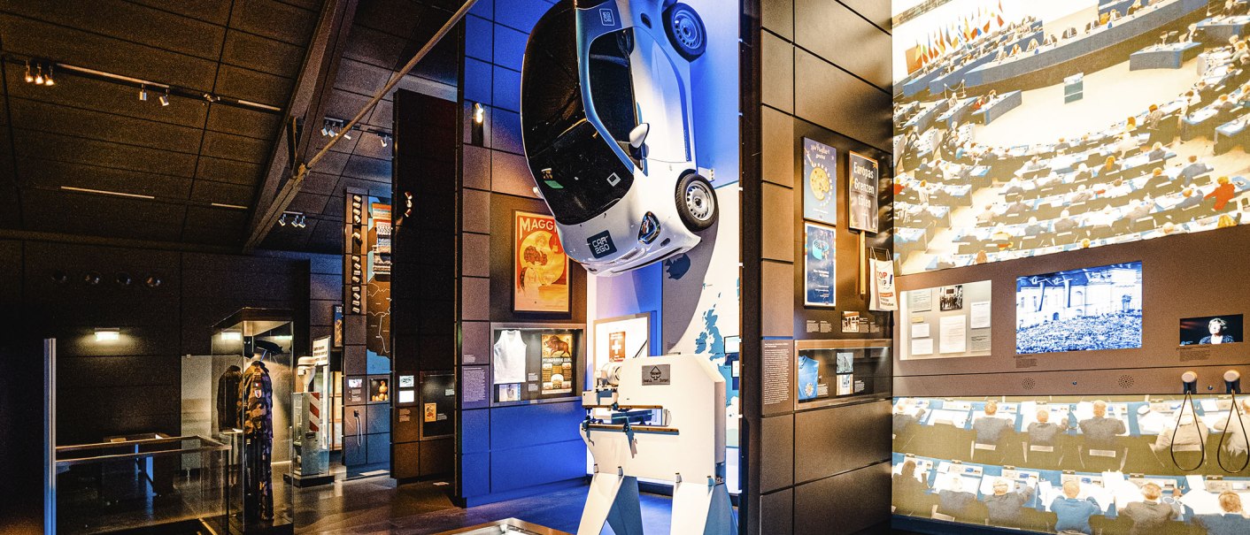 Innenansicht der Dauerausstellung im Haus der Geschichte. Ein Auto hängt an der Wand, umgeben von historischen Exponaten und Projektionen., © Stuttgart Marketing GmbH, Sarah Schmid