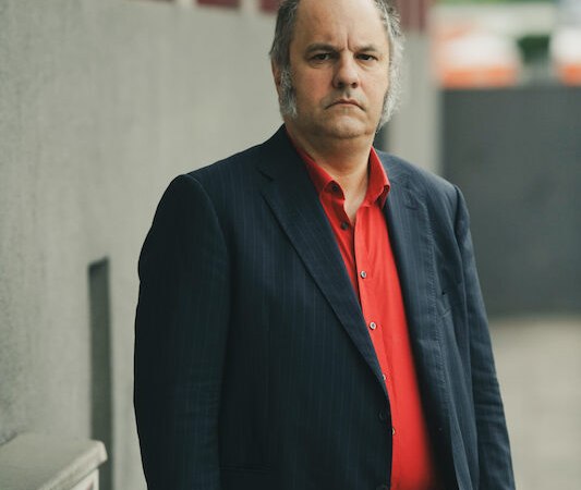 Ein Mann in einem dunklen Anzug und rotem Hemd steht ernsthaft vor einer grauen Wand im Freien., © Renitenztheater Stuttgart e.V. Ein Mann in einem dunklen Anzug und rotem Hemd steht ernsthaft vor einer grauen Wand im Freien., © Renitenztheater Stuttgart e.V.