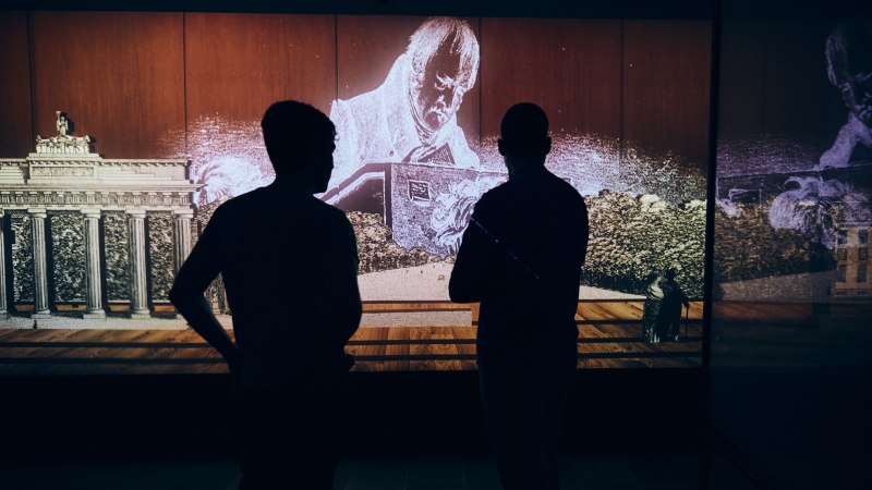 Two people stand in front of a multimedia installation showing Hegel and the Brandenburg Gate. The scene is dark and atmospherically lit., © Julia Ochs