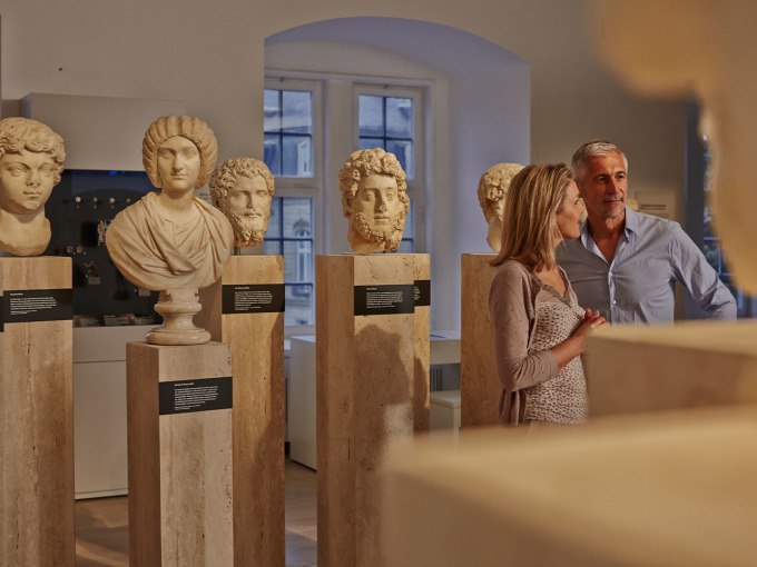 Ein Paar betrachtet antike Büsten in einem Museum. Die Skulpturen sind auf Steinsockeln ausgestellt, umgeben von weichem Licht., © Landesmuseum Württemberg Ein Paar betrachtet antike Büsten in einem Museum. Die Skulpturen sind auf Steinsockeln ausgestellt, umgeben von weichem Licht., © Landesmuseum Württemberg