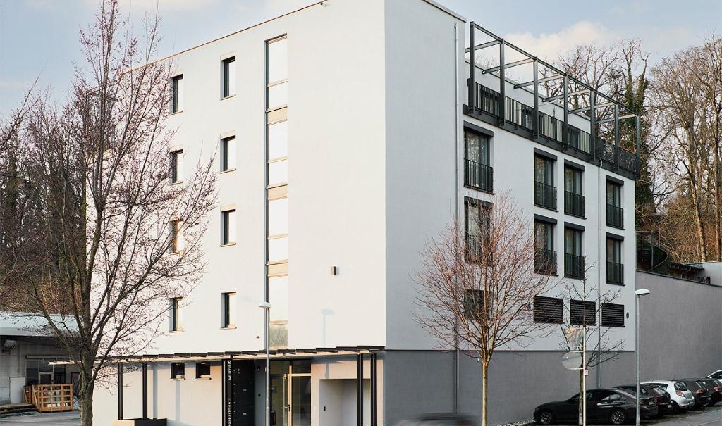 Modernes, mehrstöckiges Gebäude mit großen Fenstern und Balkonen, umgeben von Bäumen und parkenden Autos., © livisit bergapartments