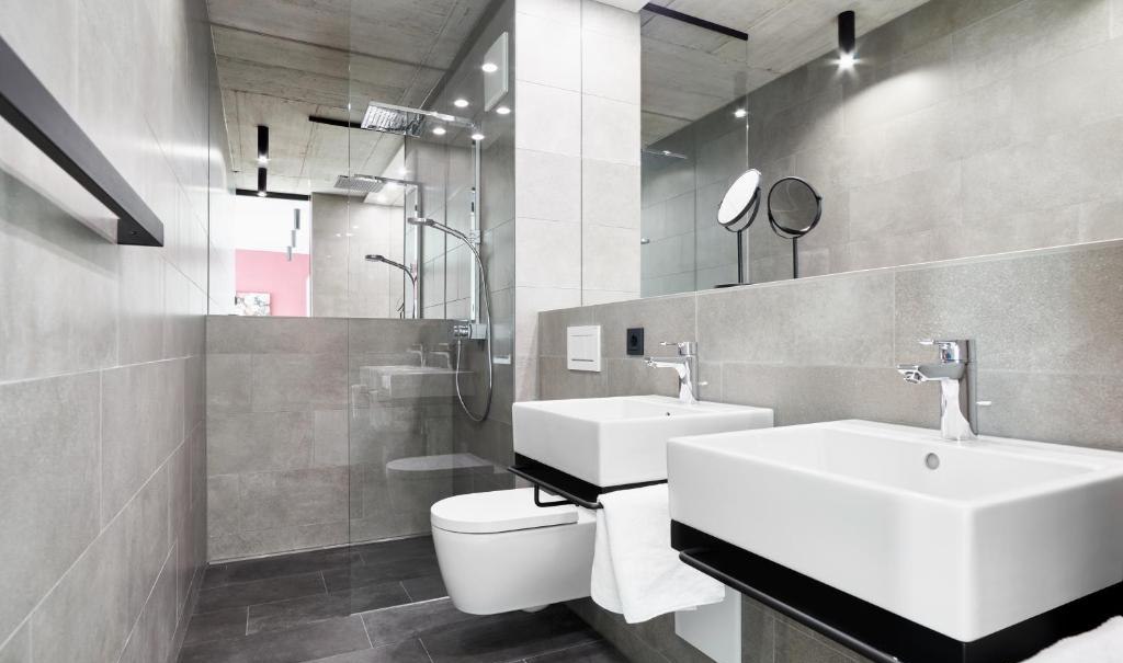 Modernes Badezimmer mit zwei Waschbecken, großer Spiegelwand, Regendusche und grauen Fliesen. Minimalistisches Design mit klaren Linien., © livisit bergapartments