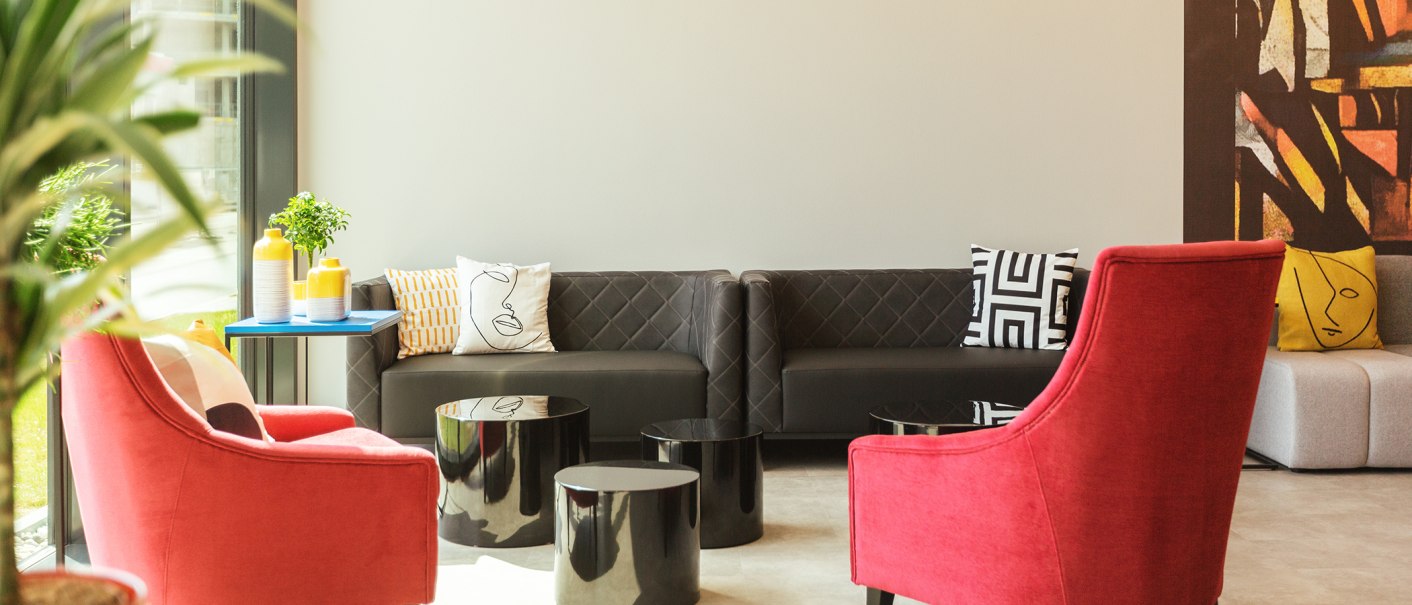 A modern lobby with red armchairs, a gray sofa, and colorful pillows. A blue table with yellow vases and plants stands in the room., © the niu Form - Novum Hospitality A modern lobby with red armchairs, a gray sofa, and colorful pillows. A blue table with yellow vases and plants stands in the room., © the niu Form - Novum Hospitality