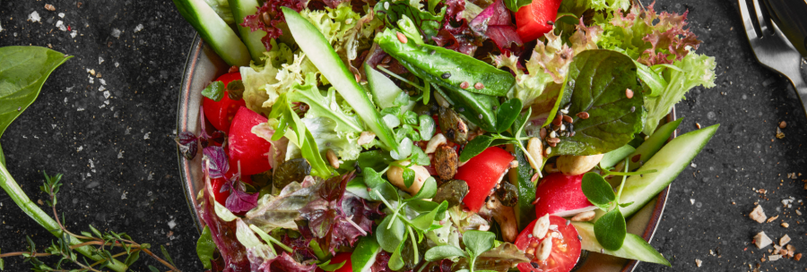 A colorful salad with tomatoes, cucumbers, lettuce leaves and seeds on a plate. Next to it are cutlery and herbs on a dark background., © MAREDO