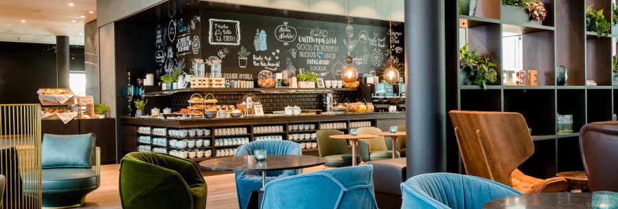 Moderne Lobby mit bunten Sesseln, Tischen und einer Kaffeebar. Die Wand ist mit Kreidezeichnungen dekoriert. Pflanzen und Regale erg&auml;nzen das Ambiente., &copy; Motel One - Stuttgart Hauptbahnhof