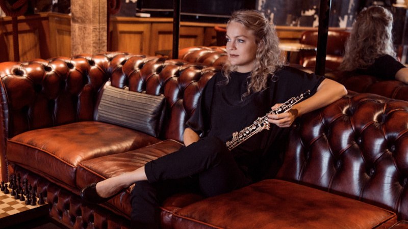 Eine Frau sitzt entspannt auf einem roten Ledersofa und hält eine Oboe. Der Raum ist gemütlich eingerichtet mit Schachbrett und Fernbedienung auf dem Tisch., © Klassische Philharmonie Bonn e.V.