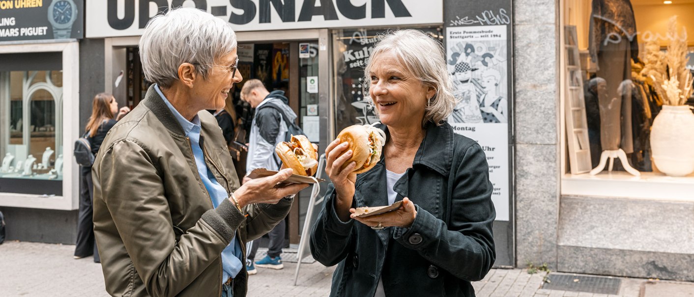 Zwei Frauen genießen Burger vor dem Imbiss 'UDO SNACK' in einer belebten Straße. Sie lachen und unterhalten sich., © Stuttgart Marketing GmbH - Sarah Schmid Zwei Frauen genießen Burger vor dem Imbiss 'UDO SNACK' in einer belebten Straße. Sie lachen und unterhalten sich., © Stuttgart Marketing GmbH - Sarah Schmid