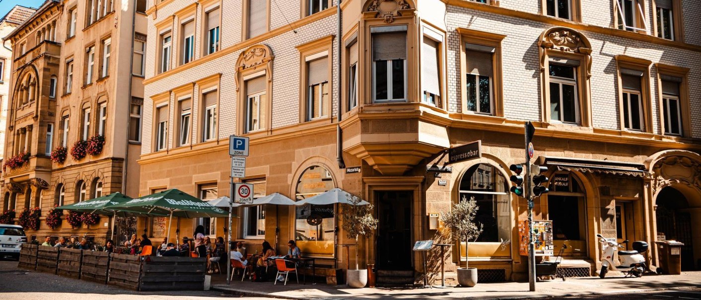Straßencafé mit Sonnenschirmen vor einem historischen Gebäude. Menschen sitzen draußen, genießen das Wetter. Verkehrsschilder und eine Vespa sind sichtbar., © Stuttgart Marketing GmbH, Sarah Schmid