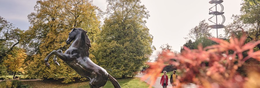 Bronze statue of an ascending horse in the Höhenpark Killesberg, surrounded by autumnal trees and a spiral-shaped observation tower in the background., © SMG, Christoph Düpper