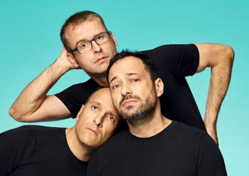 Three men in black T-shirts pose in front of a turquoise background. They look serious and are leaning close together., © Rosenau Kultur e.V. Three men in black T-shirts pose in front of a turquoise background. They look serious and are leaning close together., © Rosenau Kultur e.V.
