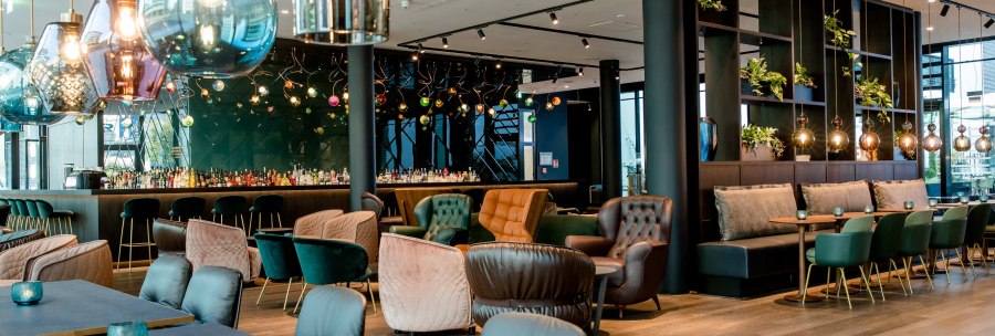 Ein modernes Restaurant mit eleganten H&auml;ngelampen, gem&uuml;tlichen Sesseln und einer Bar im Hintergrund. Pflanzen dekorieren die Regale., &copy; Motel One - Stuttgart Hauptbahnhof