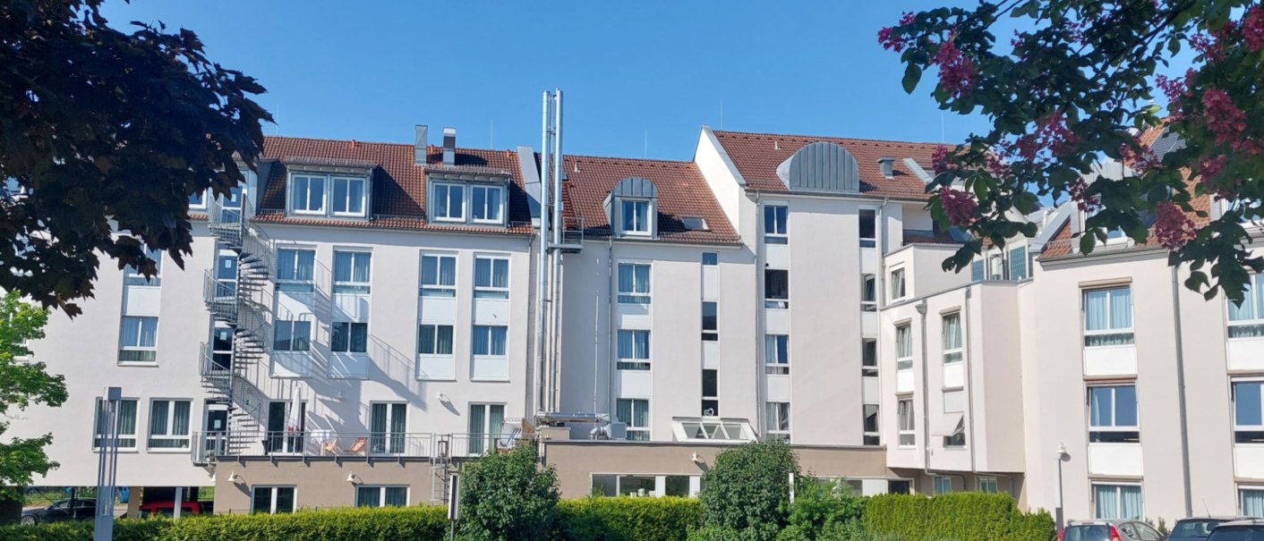 Mehrstöckiges Gebäude mit weißen Fassaden und roten Dächern, umgeben von Bäumen und einem Parkplatz. Der Himmel ist klar und blau., © Erikson Hotel Mehrstöckiges Gebäude mit weißen Fassaden und roten Dächern, umgeben von Bäumen und einem Parkplatz. Der Himmel ist klar und blau., © Erikson Hotel