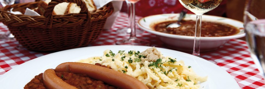 A plate of lentils, spaetzle and sausages on a red and white checkered tablecloth. A basket of bread and a glass of wine in the background., © Stuttgart-Marketing GmbH A plate of lentils, spaetzle and sausages on a red and white checkered tablecloth. A basket of bread and a glass of wine in the background., © Stuttgart-Marketing GmbH