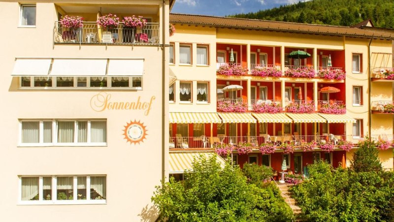 Das Hotel Sonnenhof zeigt eine Fassade mit bl&uuml;henden Balkonen und Sonnenschirmen. Der Schriftzug 'Sonnenhof' ist sichtbar. Im Hintergrund sind B&auml;ume zu sehen., &copy; Hotel Sonnenhof