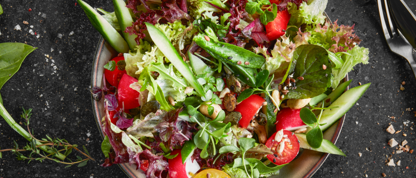 A colorful salad with tomatoes, cucumbers, lettuce leaves and seeds on a plate. Next to it are cutlery and herbs on a dark background., © MAREDO A colorful salad with tomatoes, cucumbers, lettuce leaves and seeds on a plate. Next to it are cutlery and herbs on a dark background., © MAREDO