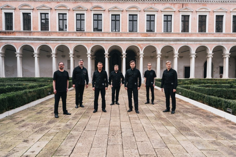 Sieben Männer in schwarzer Kleidung posieren in einem historischen Innenhof mit Arkaden und gepflegten Hecken., © Dionysos Now! Sieben Männer in schwarzer Kleidung posieren in einem historischen Innenhof mit Arkaden und gepflegten Hecken., © Dionysos Now!