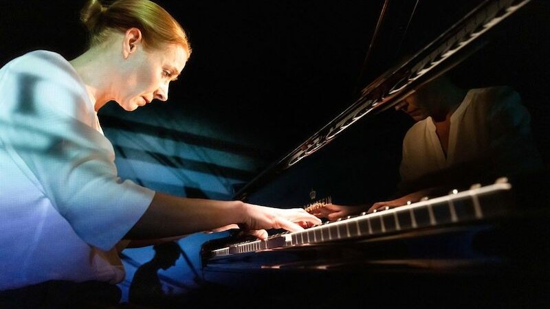 Eine Frau spielt konzentriert Klavier in einem dunklen Raum, das Licht fällt sanft auf ihr Gesicht und die Tasten., © Altes Schauspielhaus und Komödie im Marquardt e.V. Eine Frau spielt konzentriert Klavier in einem dunklen Raum, das Licht fällt sanft auf ihr Gesicht und die Tasten., © Altes Schauspielhaus und Komödie im Marquardt e.V.