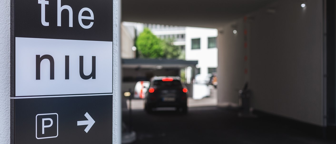 A sign with "the niu" and a parking lot symbol points to the right. A car is driving into an underground parking garage., © the niu Form - Novum Hospitality A sign with "the niu" and a parking lot symbol points to the right. A car is driving into an underground parking garage., © the niu Form - Novum Hospitality