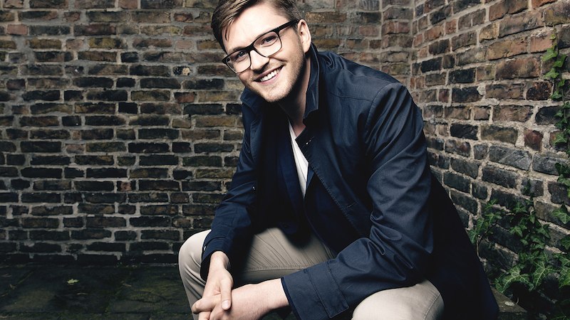 Ein Mann mit Brille und dunklem Mantel sitzt l&auml;chelnd vor einer Backsteinmauer., &copy; Internationale Hugo-Wolf-Akademie