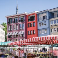 Wochenmarkt in Stuttgart mit bunten Marktständen und Gebäuden im Hintergrund. Menschen schlendern zwischen den Ständen, während die Sonne scheint., © SMG, trickytine