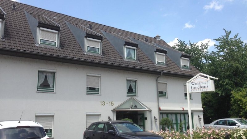 Das Aparthotel Wangener Landhaus mit grauem Dach und weißen Wänden. Vor dem Gebäude sind Autos geparkt. Ein Schild zeigt den Hotelnamen., © Aparthotel Wangener Landhaus Das Aparthotel Wangener Landhaus mit grauem Dach und weißen Wänden. Vor dem Gebäude sind Autos geparkt. Ein Schild zeigt den Hotelnamen., © Aparthotel Wangener Landhaus