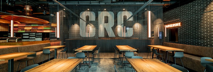 Modern café with wooden tables, chairs and hanging lamps. "CRC" on the wall, bottle racks in the background, checkroom sign illuminated., © Maks Richter