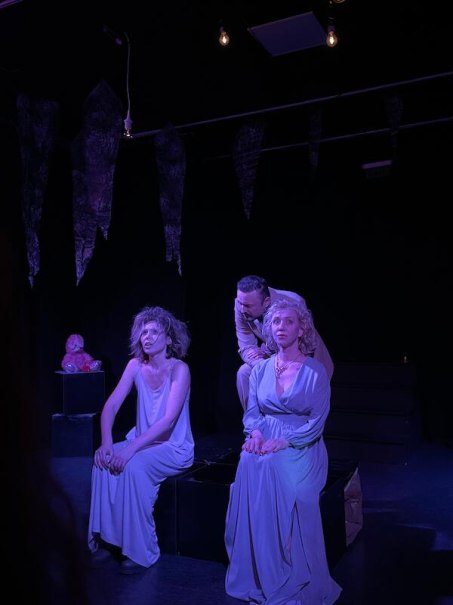 Three people in a dark room, dramatically lit in violet. Two are sitting on boxes, one is standing behind them. The scene looks theatrical., &copy; Theater Atelier