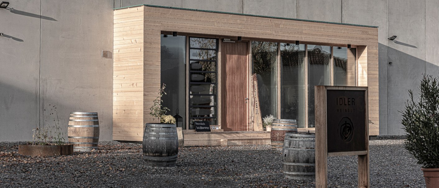 Modern entrance to the Idler winery with wooden cladding, large windows and wine barrels. A sign with the name of the winery stands in the foreground., © Weingut Idler