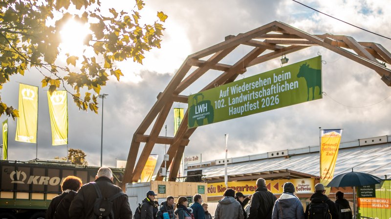 Besucher betreten das Landwirtschaftliche Hauptfest 2026 unter einem Holzbogen mit Banner. Herbstliche Atmosph&auml;re mit gelben Bl&auml;ttern und Wolken., &copy; Daniel Schneider, 2022