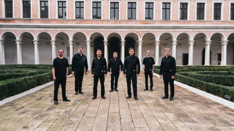 Sieben Männer in schwarzer Kleidung posieren in einem historischen Innenhof mit Arkaden und gepflegten Hecken., © Dionysos Now! Sieben Männer in schwarzer Kleidung posieren in einem historischen Innenhof mit Arkaden und gepflegten Hecken., © Dionysos Now!