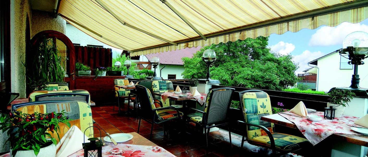 Eine Terrasse mit gedeckten Tischen, bunten Stühlen und einer gestreiften Markise. Pflanzen und Lampen schmücken den Bereich., © Hotel & Restaurant Beurener Hof Eine Terrasse mit gedeckten Tischen, bunten Stühlen und einer gestreiften Markise. Pflanzen und Lampen schmücken den Bereich., © Hotel & Restaurant Beurener Hof