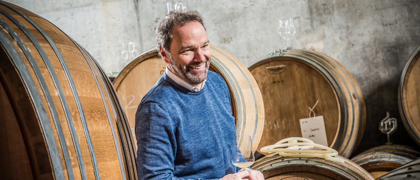 Ein Mann in einem blauen Pullover steht lächelnd vor großen Holzfässern in einem Weinkeller., © Weingut Schnaitmann