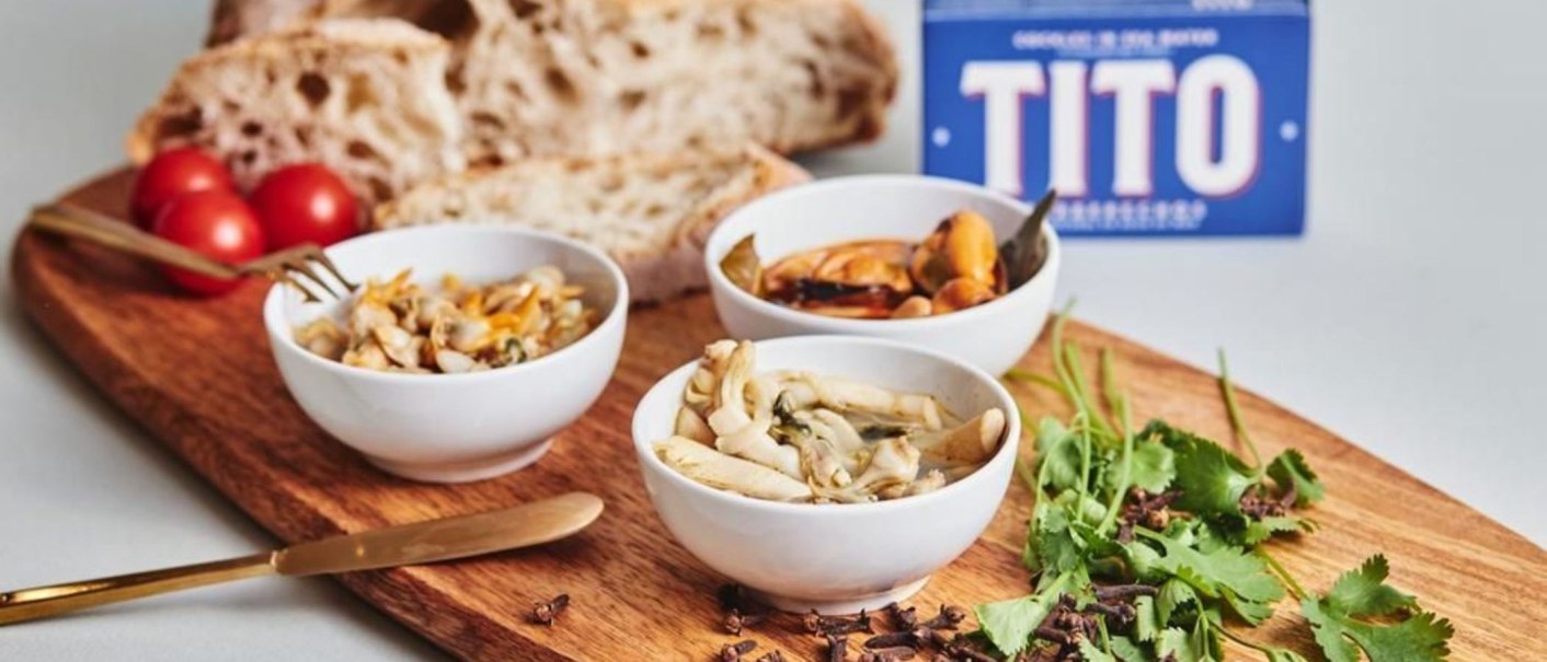 Holzbrett mit Schalen voller Meeresfrüchte, Brot, Tomaten und Kräutern. Im Hintergrund eine Verpackung mit der Aufschrift 'TITO'., © Sardinen und Feinkost, Stuttgart
