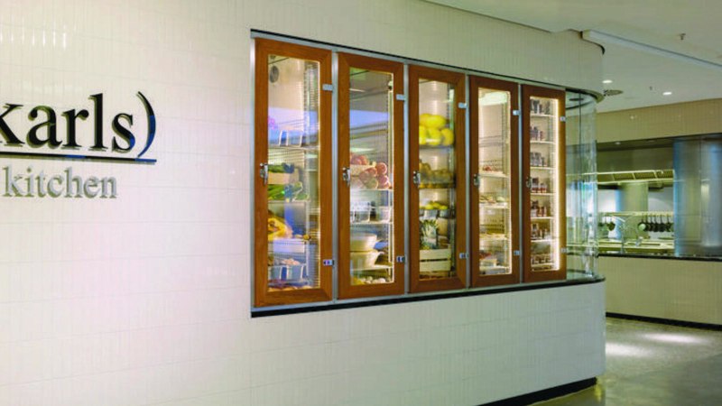Eingang zu "karls kitchen" mit beleuchteten Glasvitrinen voller Lebensmittel und Blick in die moderne Küche., © karls kitchen, Stuttgart