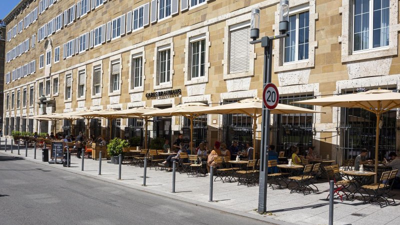 Carls Brauhaus mit Stra&szlig;encaf&eacute;, Sonnenschirmen und G&auml;sten auf einer belebten Stra&szlig;e bei sonnigem Wetter., &copy; SMG, Sarah Schmid