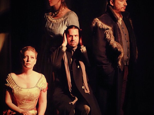 Vier Personen in historischer Kleidung posieren auf einer Bühne. Eine Frau sitzt vorne, während die anderen stehen. Die Szene ist dramatisch beleuchtet., © Theater Atelier Vier Personen in historischer Kleidung posieren auf einer Bühne. Eine Frau sitzt vorne, während die anderen stehen. Die Szene ist dramatisch beleuchtet., © Theater Atelier