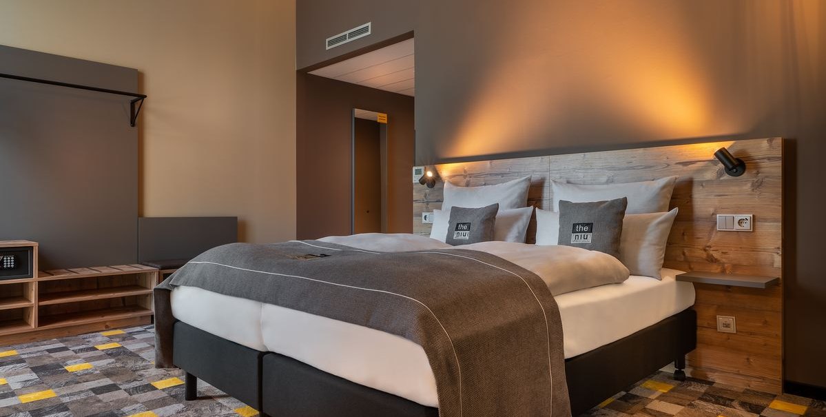 Modernes Hotelzimmer mit Doppelbett, Holzkopfteil, grauen Kissen und gemustertem Teppich in Grau und Gelb., © Novum Hospitality