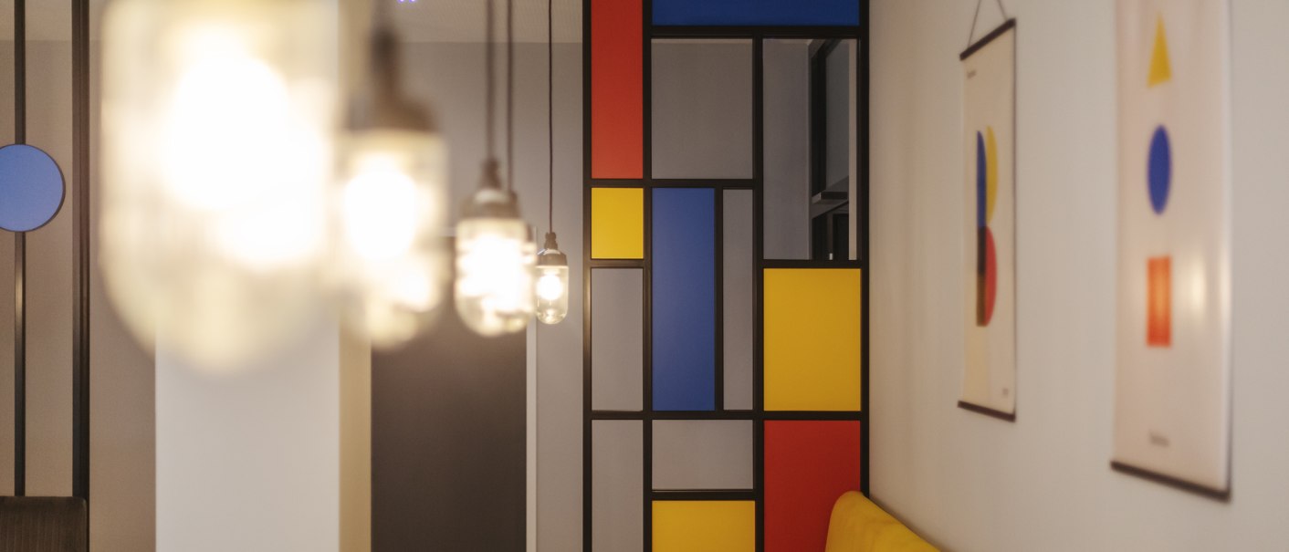 A restaurant with a geometric wall design in red, blue, and yellow. Pendant lights in the foreground, modern art on the walls., © the niu Form - Novum Hospitality A restaurant with a geometric wall design in red, blue, and yellow. Pendant lights in the foreground, modern art on the walls., © the niu Form - Novum Hospitality
