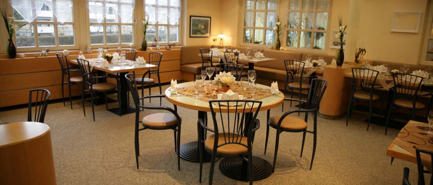 Ein elegantes Restaurant mit gedeckten Tischen, Stühlen und dekorativen Blumen. Große Fenster lassen viel Licht herein., © ateckhotel