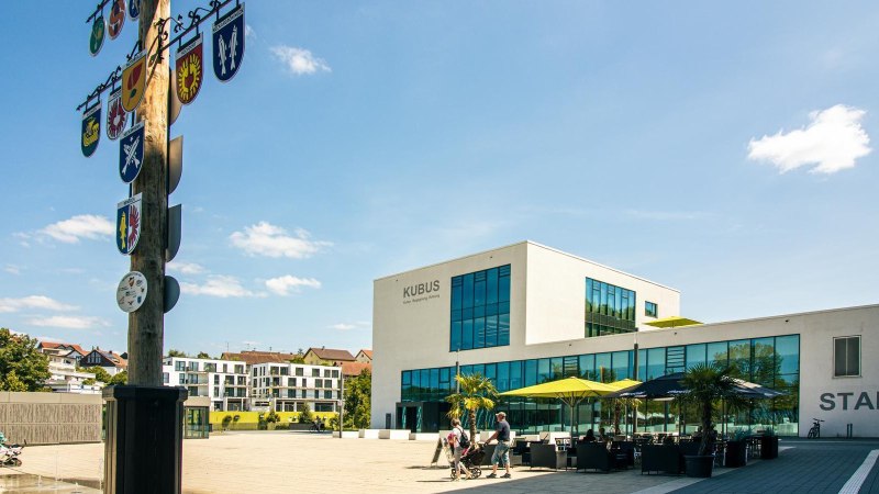 Modern market square in Remseck with the 'KUBUS' building, a maypole with a coat of arms and people out for a walk. Sunny day with blue sky., © Stuttgart-Marketing GmbH, Sarah Schmid Modern market square in Remseck with the 'KUBUS' building, a maypole with a coat of arms and people out for a walk. Sunny day with blue sky., © Stuttgart-Marketing GmbH, Sarah Schmid