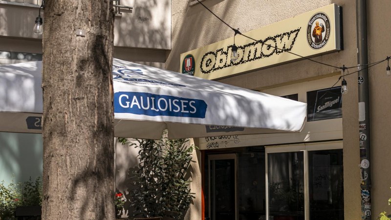 Eingang eines Caf&eacute;s namens Oblamow mit Gauloises-Sonnenschirm und Pflanzen. Au&szlig;enbeleuchtung und Schilder von Franziskaner und San Miguel sichtbar., &copy; SMG, Sarah Schmid