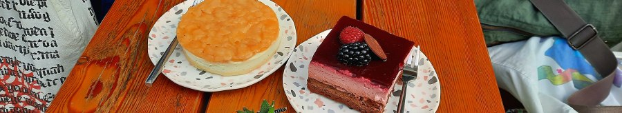 Two pieces of cake on plates with forks on a wooden table, next to a small plant., © SMG