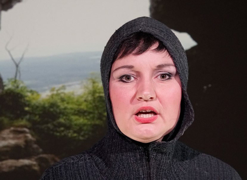 Eine Frau mit dunkler Kapuze und starkem Make-up steht vor einem Landschaftshintergrund mit B&auml;umen und Felsen., &copy; Einfach sagenhaft-Ellen Schubert