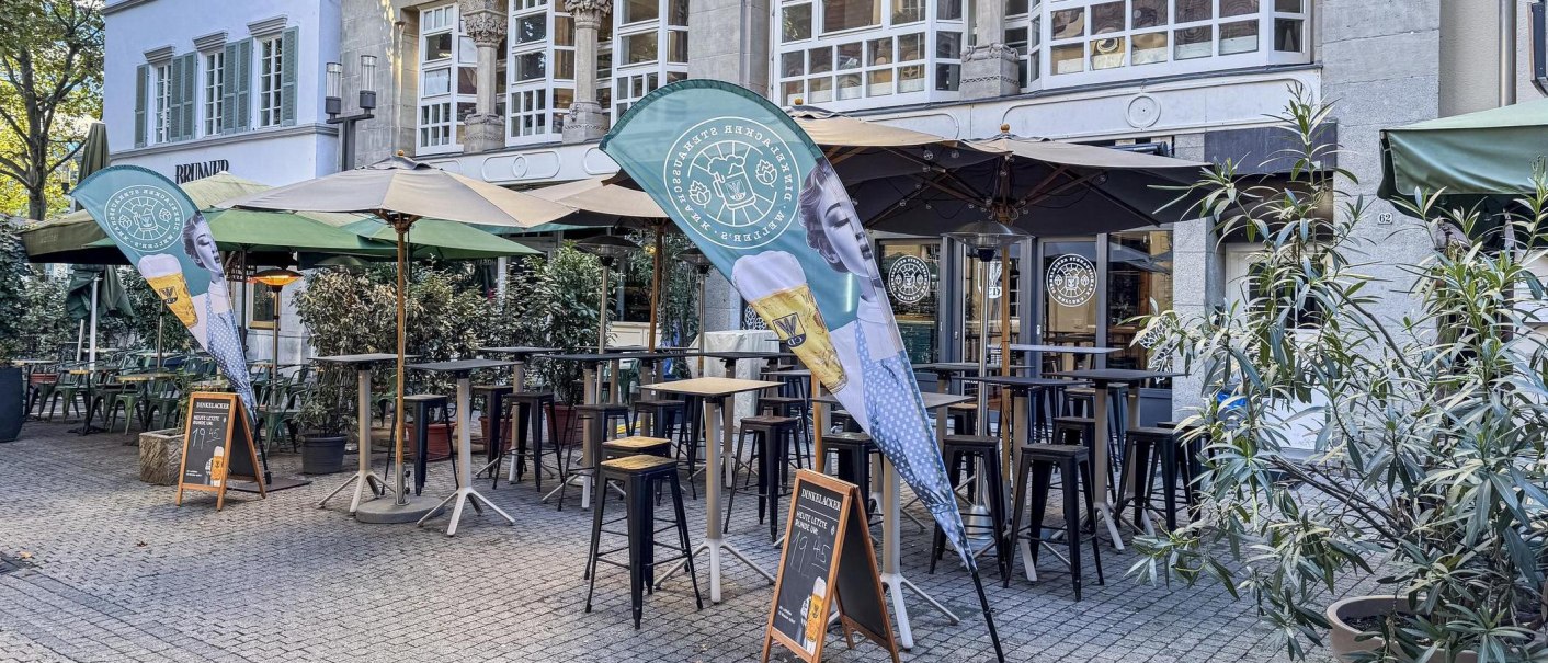 Außenbereich des Dinkelacker-Stehausschanks mit Stehtischen, Sonnenschirmen und Bierfahnen vor einem historischen Gebäude., © SMG, Ellen Haeffner Außenbereich des Dinkelacker-Stehausschanks mit Stehtischen, Sonnenschirmen und Bierfahnen vor einem historischen Gebäude., © SMG, Ellen Haeffner