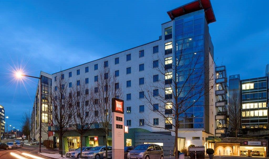 Außenansicht eines mehrstöckigen Ibis Hotels bei Dämmerung, beleuchtete Fenster, geparkte Autos, Straßenlaterne und blauer Himmel., © TOMAS Außenansicht eines mehrstöckigen Ibis Hotels bei Dämmerung, beleuchtete Fenster, geparkte Autos, Straßenlaterne und blauer Himmel., © TOMAS