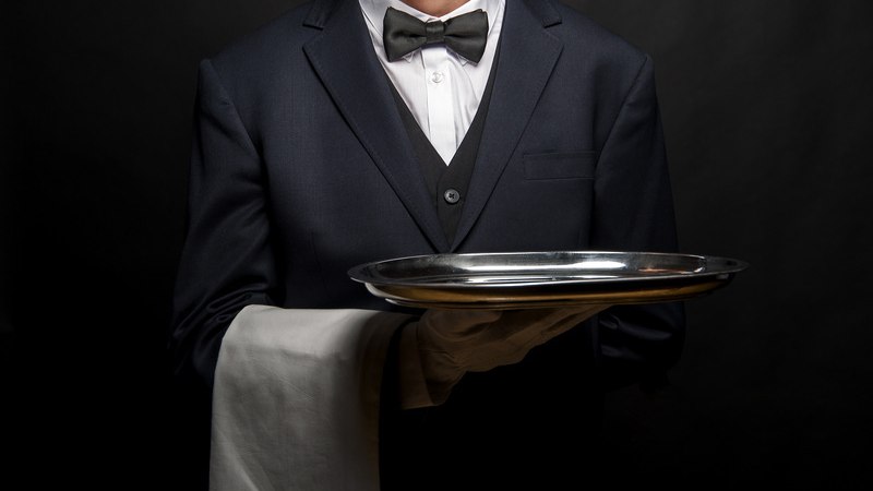 Ein Kellner in Anzug und Fliege präsentiert ein silbernes Tablett, vor einem dunklen Hintergrund. Er trägt ein weißes Tuch über dem Arm., © engesser marketing GmbH Ein Kellner in Anzug und Fliege präsentiert ein silbernes Tablett, vor einem dunklen Hintergrund. Er trägt ein weißes Tuch über dem Arm., © engesser marketing GmbH