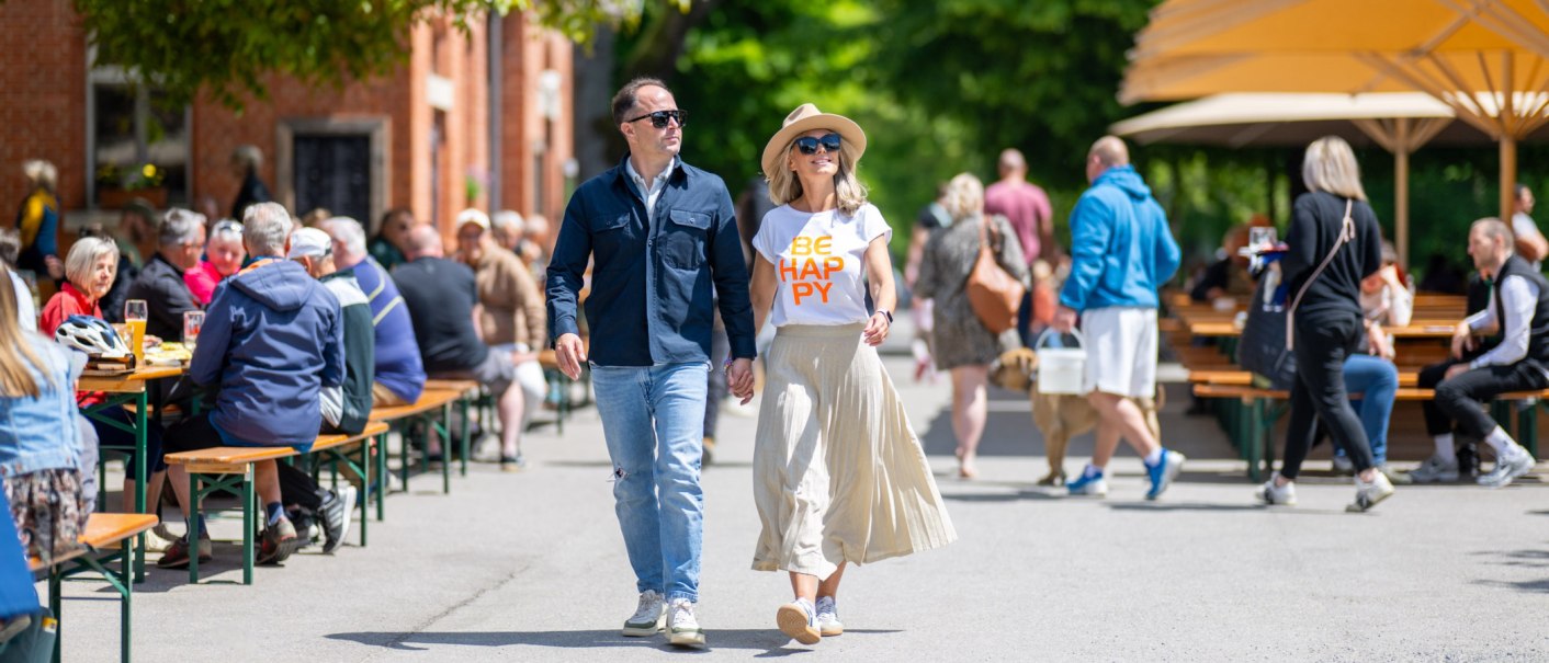 A couple is strolling through a bustling beer garden. They are wearing casual clothes and sunglasses. In the background, people are sitting at tables under umbrellas., © Katzenbacher Hof A couple is strolling through a bustling beer garden. They are wearing casual clothes and sunglasses. In the background, people are sitting at tables under umbrellas., © Katzenbacher Hof