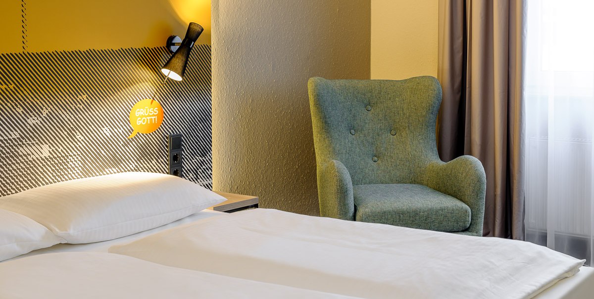 Hotelzimmer mit einem Bett, einem blauen Sessel und einer gelben Wand mit "Grüß Gott!"-Schriftzug., © TOMAS