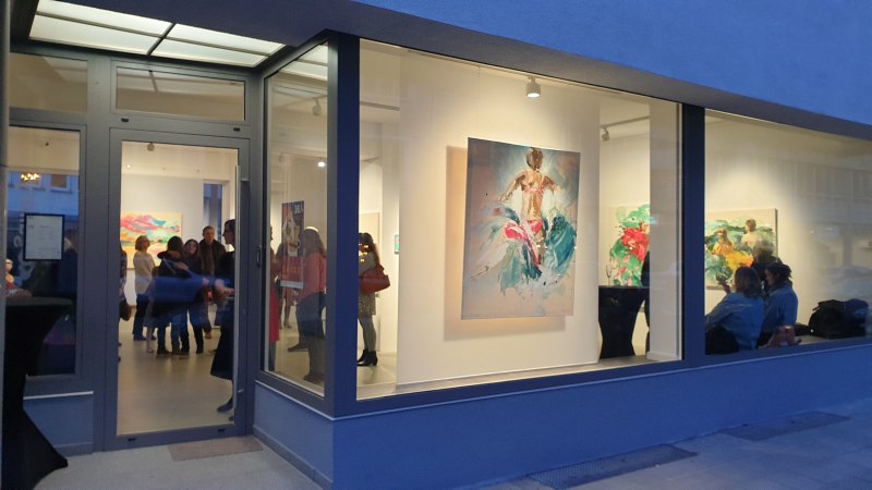 Eine Galerie mit großen Fenstern zeigt bunte Gemälde. Menschen stehen und betrachten die Kunstwerke im Inneren., © EXOgallery Contemporary and New Media Art
