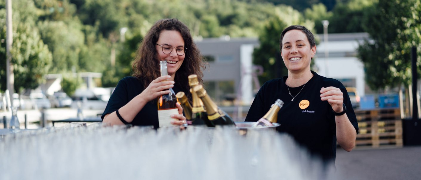 Two people are laughing and holding bottles of wine. They are standing outside, surrounded by trees and buildings in the background., © Bernhard Kahrmann Two people are laughing and holding bottles of wine. They are standing outside, surrounded by trees and buildings in the background., © Bernhard Kahrmann